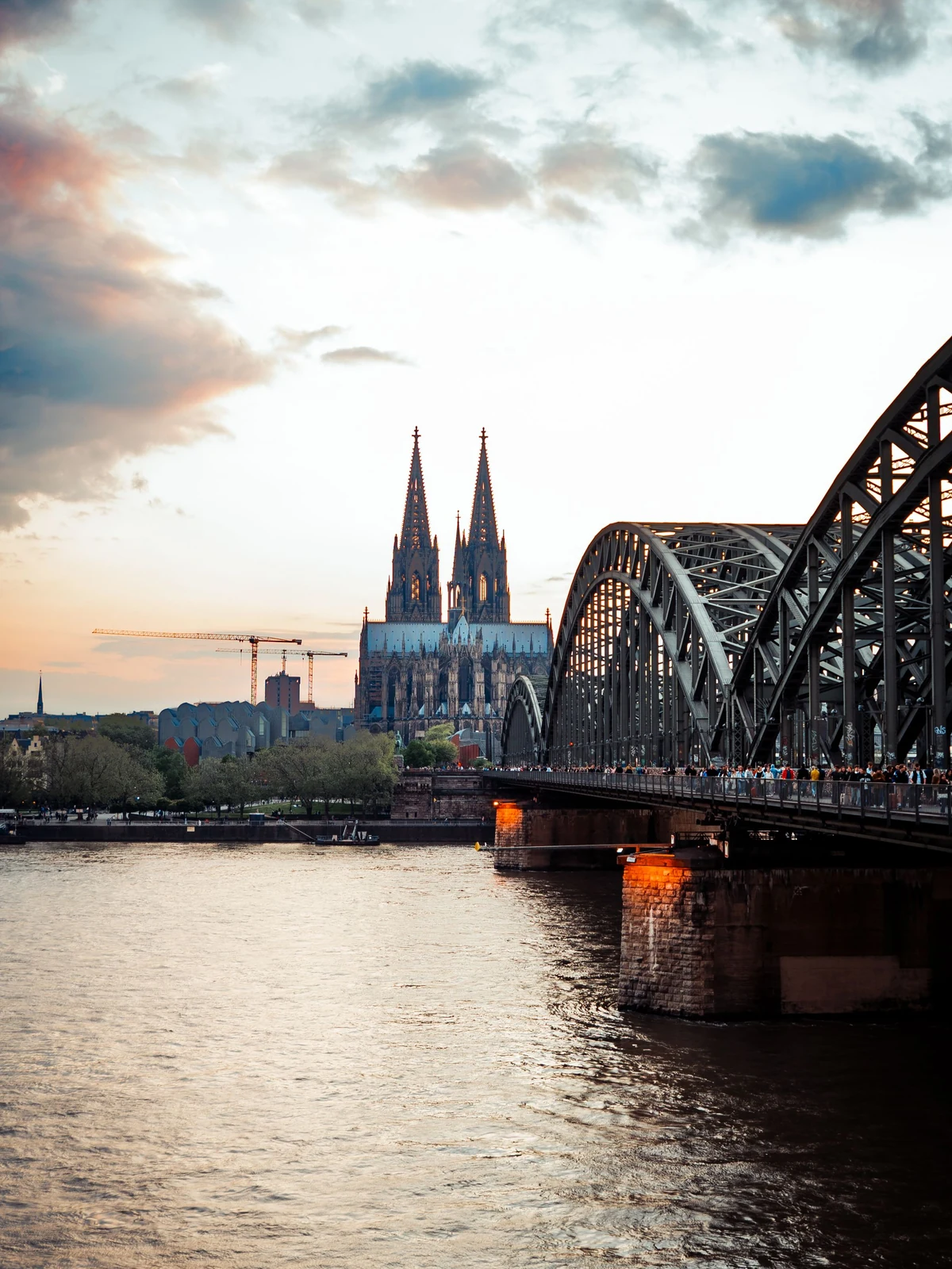 Cologne Cathedral and Hohenzollern railway bridge beside the Rhine at sunset.