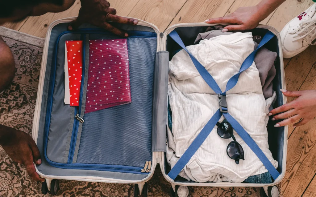 Hands packing folded clothing into an open suitcase on a wooden floor.